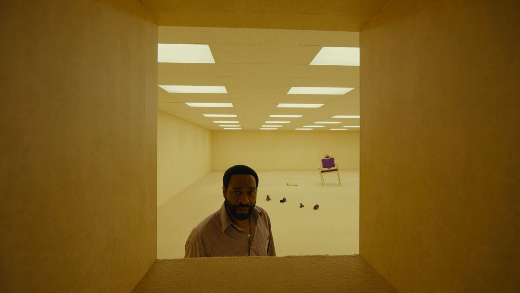 The protagonist of the movie Backrooms, a black middle-aged man with short hair and a full beard, gazes through a windowless wall opening into an empty, yellow-toned room where furniture appears to sink into the carpet.