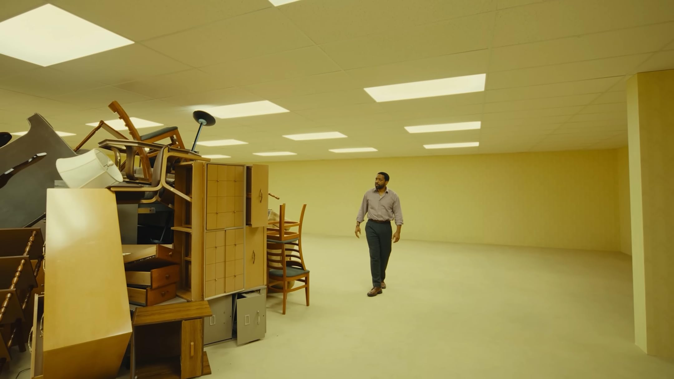 The protagonist of the movie Backrooms approaches a pile of loosely stacked furniture (chairs, sideboards, a file cabinet) at the center of an office-like empty room.